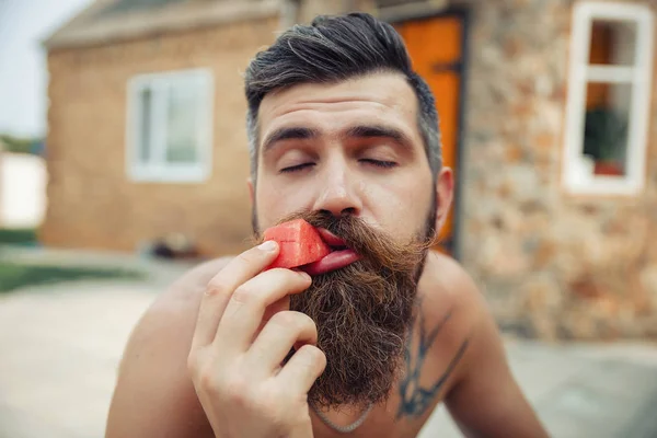 Un homme avec une coiffure élégante, une longue barbe et un tatouage avec un grand plaisir mange pastèque — Photo