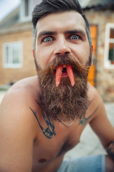 Um homem com um penteado elegante, uma longa barba e uma tatuagem com grande prazer come melancia — Fotografia de Stock