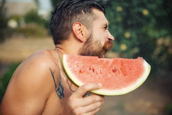 Bir adam şık bir saç modeli, uzun sakallı ve büyük bir zevk ile dövme karpuz yiyor