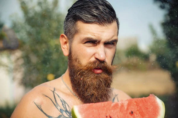Bir adam şık bir saç modeli, uzun sakallı ve büyük bir zevk ile dövme karpuz yiyor