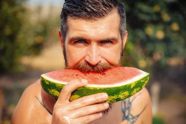 Un homme avec une coiffure élégante, une longue barbe et un tatouage avec un grand plaisir mange pastèque — Photo