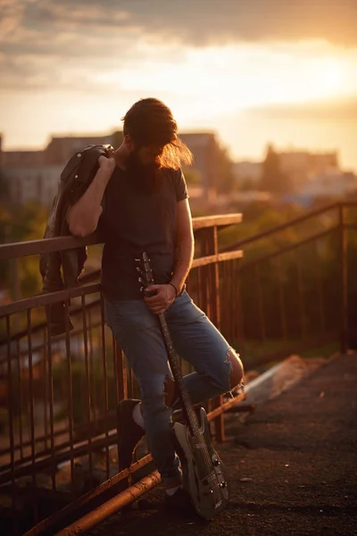 Elegantemente vestido hombre guapo con una larga barba, chaqueta de cuero marrón y jeans ligeros sosteniendo una guitarra y de pie en un puente — Foto de Stock