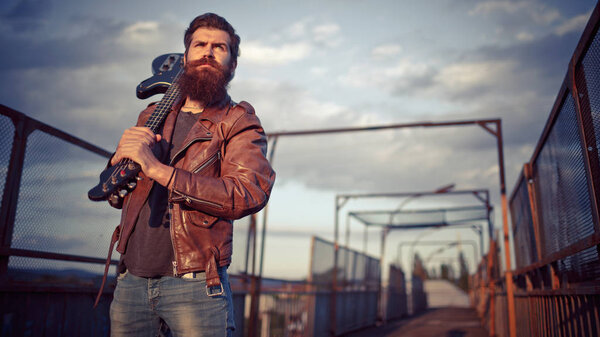Стильно одетый красивый мужчина с длинной бородой, коричневой кожаной курткой и легкими джинсами, держащий гитару и стоящий на мосту
