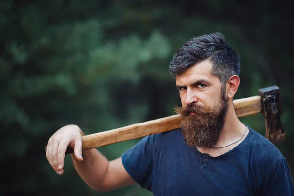 Homem barbudo elegante em uma camiseta escura, com uma cabeça na cabeça segurando em suas mãos um machado de madeira na floresta — Fotografia de Stock