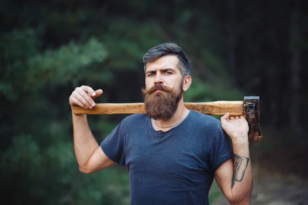 Homem barbudo elegante em uma camiseta escura, com uma cabeça na cabeça segurando em suas mãos um machado de madeira na floresta — Fotografia de Stock