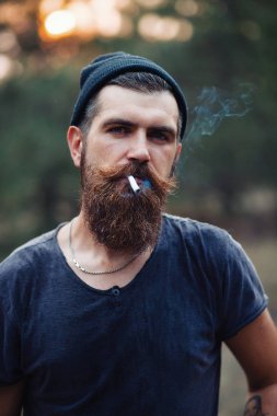 Uzun sakallı ve trendy kıyafetler şık bir oduncu bir sigara içiyor ve bir balta ormana elinde tutan