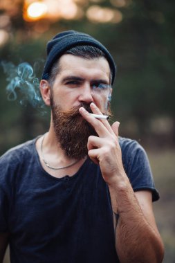 Uzun sakallı ve trendy kıyafetler şık bir oduncu bir sigara içiyor ve bir balta ormana elinde tutan