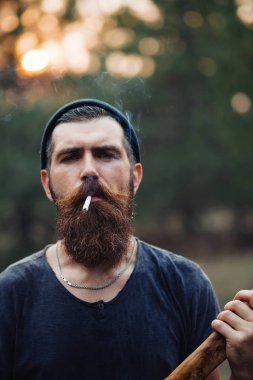Uzun sakallı ve trendy kıyafetler şık bir oduncu bir sigara içiyor ve bir balta ormana elinde tutan
