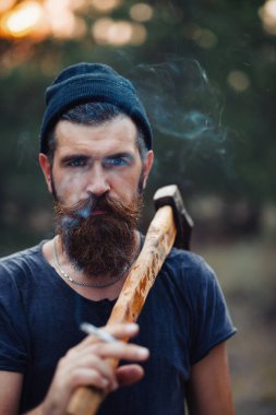 Uzun sakallı ve trendy kıyafetler şık bir oduncu bir sigara içiyor ve bir balta ormana elinde tutan