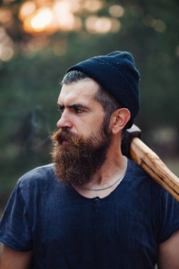 Uzun sakallı ve trendy kıyafetler şık bir oduncu bir sigara içiyor ve bir balta ormana elinde tutan