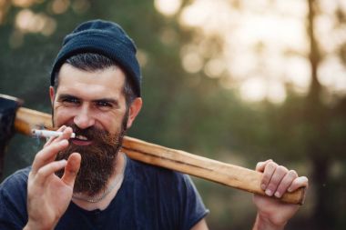 Uzun sakallı ve trendy kıyafetler şık bir oduncu bir sigara içiyor ve bir balta ormana elinde tutan
