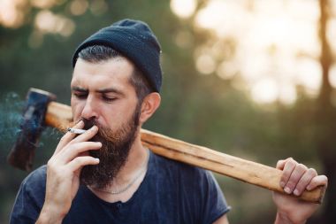 Uzun sakallı ve trendy kıyafetler şık bir oduncu bir sigara içiyor ve bir balta ormana elinde tutan