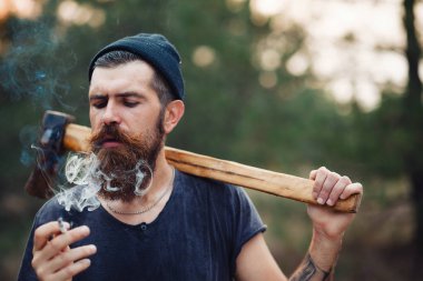 Uzun sakallı ve trendy kıyafetler şık bir oduncu bir sigara içiyor ve bir balta ormana elinde tutan