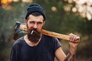 Uzun sakallı ve trendy kıyafetler şık bir oduncu bir sigara içiyor ve bir balta ormana elinde tutan