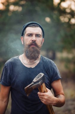 Uzun sakallı ve trendy kıyafetler şık bir oduncu bir sigara içiyor ve bir balta ormana elinde tutan