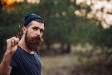 Siyah tişörtlü şık sakallı adam, kafasında kafasıyla ormanda tahta bir balta tutuyor.