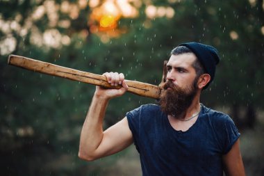 Siyah tişörtlü şık sakallı adam, kafasında kafasıyla ormanda tahta bir balta tutuyor.