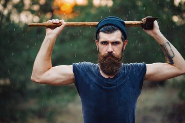 Siyah tişörtlü şık sakallı adam, kafasında kafasıyla ormanda tahta bir balta tutuyor.
