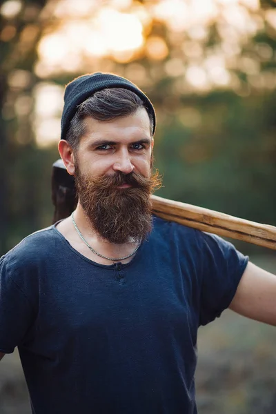 Stylový vousatý muž v tmavém tričku s hlavou na hlavě a dřevěnou sekerou v lese. — Stock fotografie