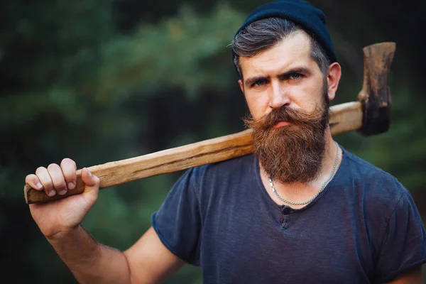 身穿深色T恤，留着时髦胡子的男人，头上顶着头，手里拿着一把木头斧子，躺在树林里 — 图库照片