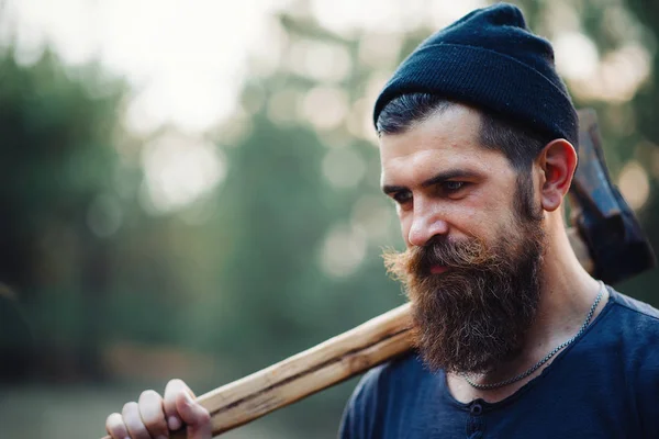 Stylish bearded man in a dark T-shirt, with a head on his head holding in his hands a wooden ax in the woods — Stock Photo, Image