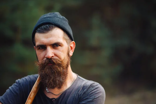 Homem barbudo elegante em uma camiseta escura, com uma cabeça na cabeça segurando em suas mãos um machado de madeira na floresta — Fotografia de Stock