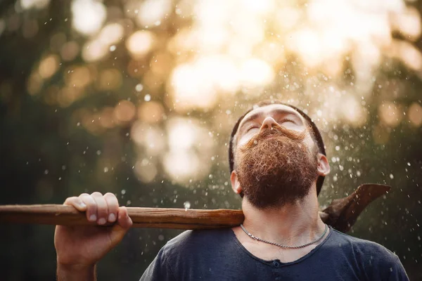 Elegante hombre barbudo con una camiseta oscura, con una cabeza en la cabeza sosteniendo en sus manos un hacha de madera en el bosque — Foto de Stock