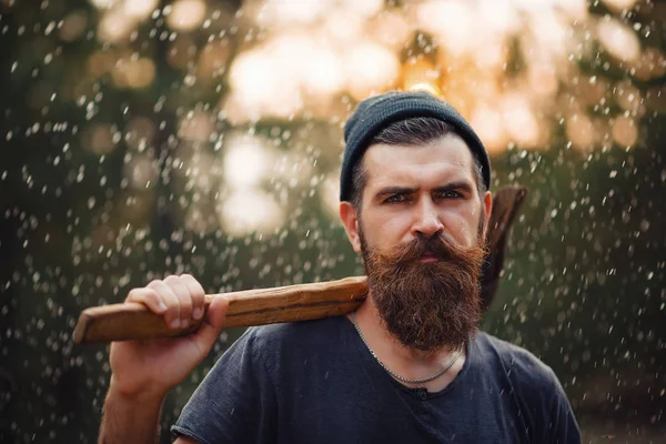 Elegante hombre barbudo con una camiseta oscura, con una cabeza en la cabeza sosteniendo en sus manos un hacha de madera en el bosque — Foto de Stock