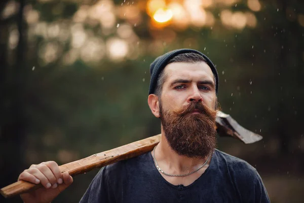 Homem barbudo elegante em uma camiseta escura, com uma cabeça na cabeça segurando em suas mãos um machado de madeira na floresta — Fotografia de Stock