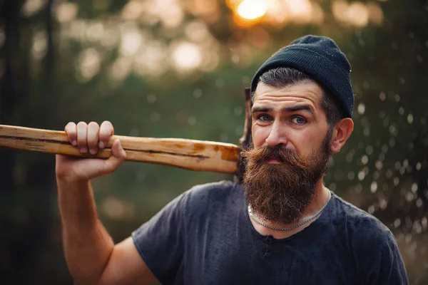 Homem barbudo elegante em uma camiseta escura, com uma cabeça na cabeça segurando em suas mãos um machado de madeira na floresta — Fotografia de Stock