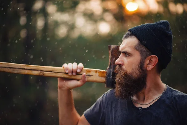 Elegante hombre barbudo con una camiseta oscura, con una cabeza en la cabeza sosteniendo en sus manos un hacha de madera en el bosque — Foto de Stock