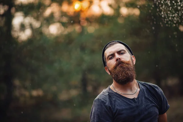 Homme barbu élégant dans un T-shirt sombre, avec une tête sur la tête tenant dans ses mains une hache en bois dans les bois — Photo