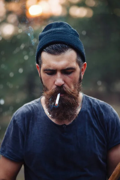 Un bûcheron élégant avec une longue barbe et des vêtements à la mode fume une cigarette et tient une hache dans les mains des bois — Photo