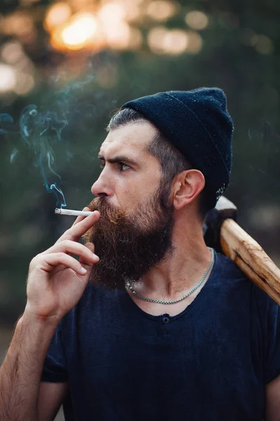 Uzun sakallı ve trendy kıyafetler şık bir oduncu bir sigara içiyor ve bir balta ormana elinde tutan