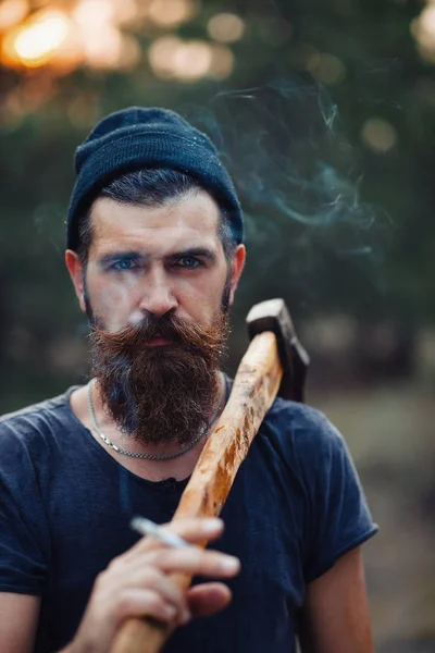 Ένα κομψό ξυλοκόπος με μακριά γενειάδα και καθιερώνοντα τη μόδα ενδύματα καπνίζει ένα τσιγάρο και κατέχει ένα τσεκούρι στα χέρια των ξύλων — Φωτογραφία Αρχείου