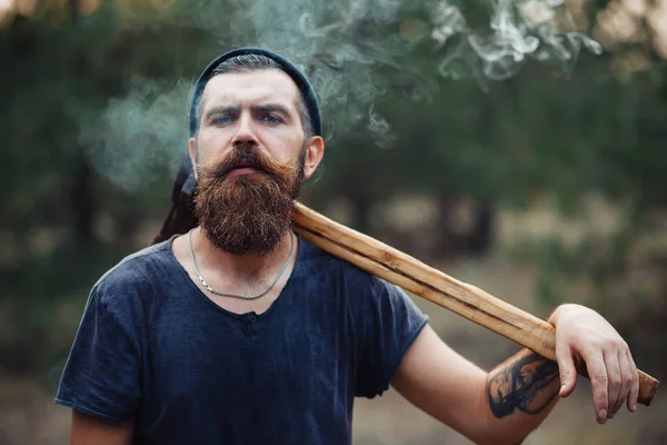 Стильный дровосек с длинной бородой и в модной одежде курит сигарету и держит топор в руках в лесу. — стоковое фото
