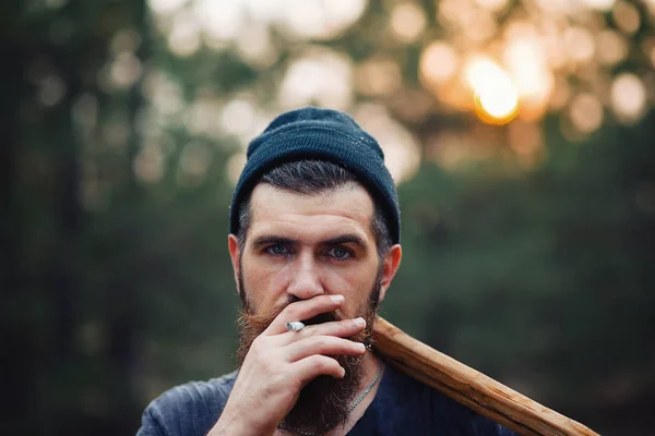 Стильный дровосек с длинной бородой и в модной одежде курит сигарету и держит топор в руках в лесу. — стоковое фото