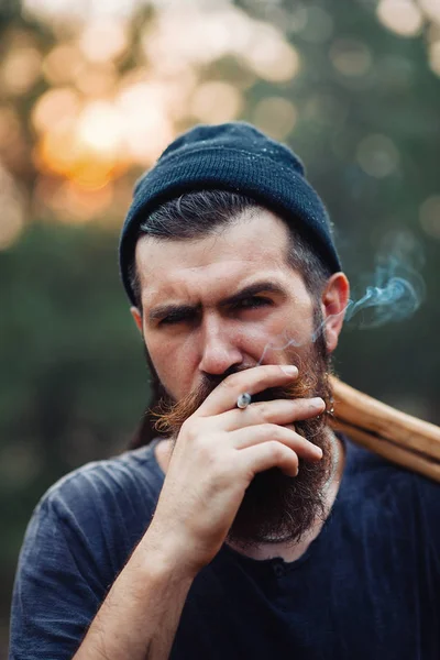 Un elegante leñador con barba larga y ropa de moda fuma un cigarrillo y sostiene un hacha en las manos del bosque —  Fotos de Stock