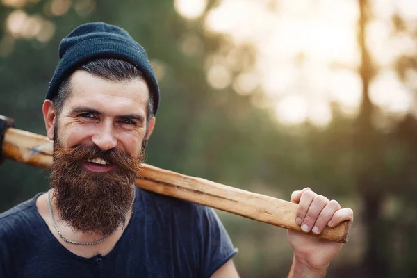 Elegante hombre barbudo con una camiseta oscura, con una cabeza en la cabeza sosteniendo en sus manos un hacha de madera en el bosque — Foto de Stock