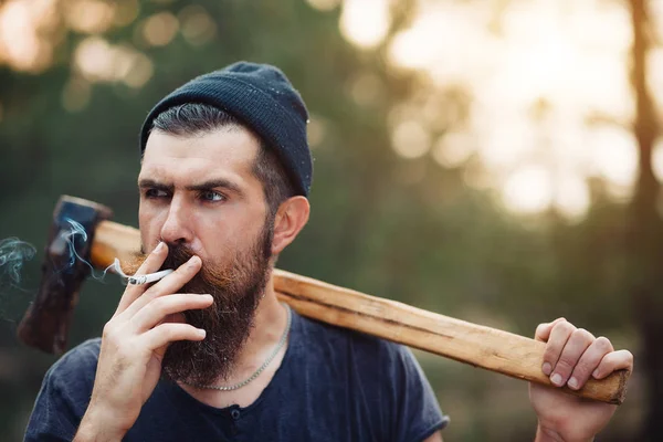 Un bûcheron élégant avec une longue barbe et des vêtements à la mode fume une cigarette et tient une hache dans les mains des bois — Photo
