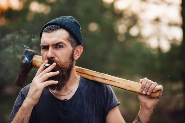 Un elegante leñador con barba larga y ropa de moda fuma un cigarrillo y sostiene un hacha en las manos del bosque — Foto de Stock