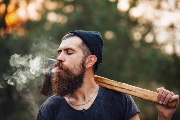 Stylowe drwal z długą brodą i w modnych ubrań pali papierosa i trzyma siekierę w rękach lasu — Zdjęcie stockowe