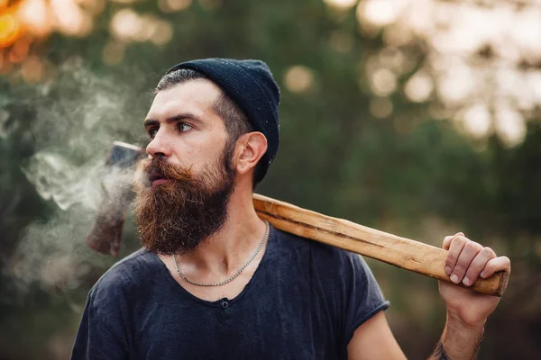 Un elegante leñador con barba larga y ropa de moda fuma un cigarrillo y sostiene un hacha en las manos del bosque — Foto de Stock