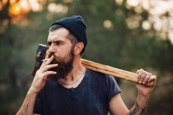 Um lenhador elegante com uma longa barba e em roupas da moda fuma um cigarro e segura um machado nas mãos da floresta — Fotografia de Stock