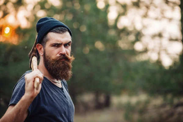 Elegante hombre barbudo con una camiseta oscura, con una cabeza en la cabeza sosteniendo en sus manos un hacha de madera en el bosque — Foto de Stock