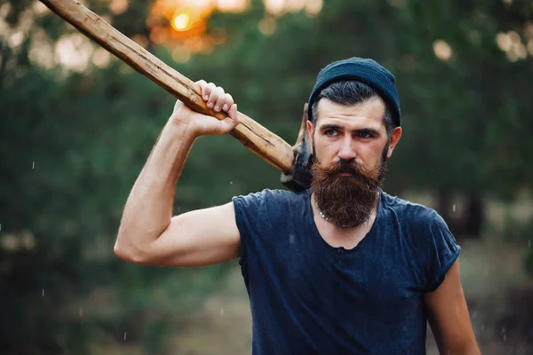 Stylowy brodaty mężczyzna w ciemnej koszulce, z głową na głowie trzymającą w dłoniach drewnianą siekierę w lesie — Zdjęcie stockowe