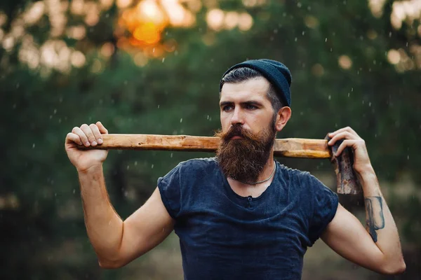 Stylish bearded man in a dark T-shirt, with a head on his head holding in his hands a wooden ax in the woods