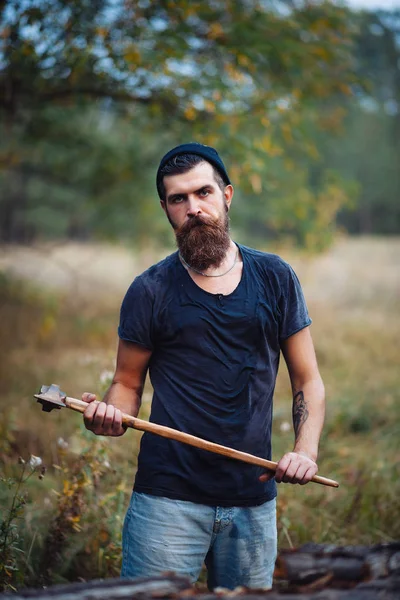 Homem barbudo elegante em uma camiseta escura, com uma cabeça na cabeça segurando em suas mãos um machado de madeira na floresta — Fotografia de Stock