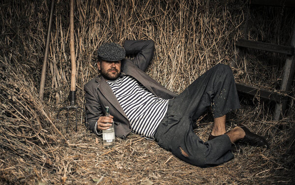 A cheerful peasant in old clothes with a bottle of alcohol in his hands lays on a dry grass in a shed