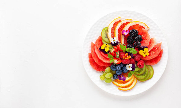 Delicious healthy salad of fresh fruits, berries and edible flowers on white plate. Clean eating. Top view.